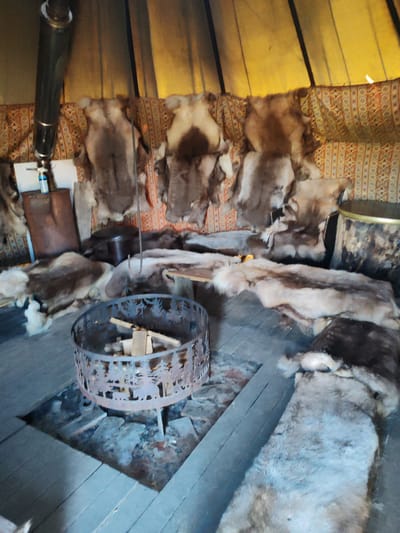 The image depicts the interior of a dwelling, likely a traditional tent or yurt, with wooden floors and fabric walls. Various animal pelts are draped over furniture and hung on the walls, suggesting a rustic or indigenous setting. A metal fire pit, containing firewood, is positioned on the floor in the foreground. A tall metal pipe, possibly a stovepipe, extends upwards on the left side of the frame. The overall impression is one of a lived-in, primitive shelter. There are no people or specific identifiable landmarks to ascertain a precise location within Lukaši, Russia, beyond it being an interior space.