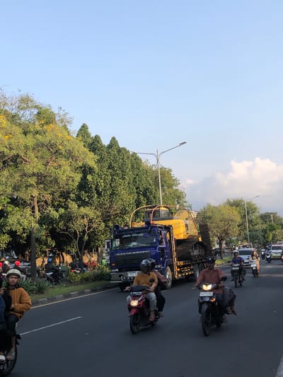This image captures a street scene in Gelam, East Java, likely during the daytime due to the clear sky and bright lighting. The activity depicts a traffic scene with several vehicles and people. The foreground shows a line of motorcycles parked and some people on them, with one individual in a yellow jacket wearing a helmet looking towards the camera. A blue truck with "NISSAN DIESEL" visible on its side is also present, along with a yellow construction vehicle. The background features lush green trees, some with yellow flowers, and a bright blue sky with scattered clouds. The overall mood appears to be casual, with people going about their day in a moderately busy urban or suburban environment. There are no strong indications of a specific event, but rather a snapshot of everyday traffic in the region.