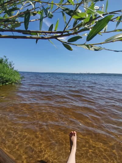 The image shows a view from a body of water, likely a lake or river, on a sunny day. In the foreground, the camera perspective is low, showing the surface of the water with light reflecting off it. A person's left foot and lower leg are visible, submerged in the shallow, clear water. The foot is positioned towards the bottom right of the frame. The water is a warm, amber color, indicating a sandy or silty bottom, which is visible through the clear water.

In the middle ground, the water extends outwards, with gentle ripples on its surface. To the left, a lush green bush or tree is growing at the water's edge. In the distance, across the water, a hazy shoreline is visible with trees and a few man-made structures, including what appears to be a factory or industrial complex with tall chimneys. The sky above is a clear, bright blue.

The image is framed from below by branches and leaves of a tree in the upper part of the frame, suggesting the viewer is seated or lying down near the water's edge, under the shade of a tree. The overall atmosphere is peaceful and summery, implying a recreational setting. This location could be a natural area adjacent to the city of Pyatyy Kilometr in Russia, perhaps a popular spot for swimming or relaxing by the water.