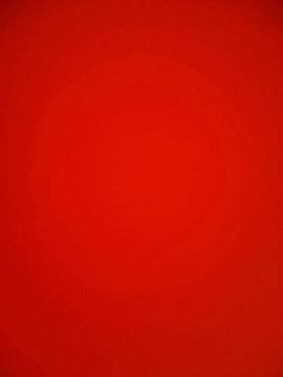 The image presents a solid, uniformly red surface. The color appears consistent throughout, with subtle gradations suggesting a slight play of light. A textured pattern is visible in the paint surface.