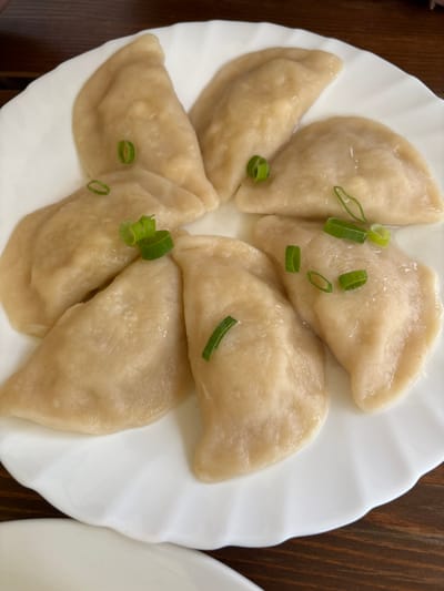 Seven light-colored pierogi are centrally arranged on a white, scalloped-edge ceramic plate. Each pierog is garnished with small, diagonally sliced pieces of green onion, appearing moist and freshly prepared. The plate is positioned on a dark brown wooden surface, likely a dining table. A partial view of another white dish is visible in the bottom-left corner. The scene depicts a served meal in an indoor environment. No individuals are present. The exact location within Wojnowo, Poland, is not discernible from the visual information provided.