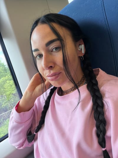 This is a portrait of a young woman indoors, possibly on a train, given the blue patterned seat and window view. She has long, dark hair styled in two braids, and is wearing a pink sweatshirt and a black choker necklace. She also has white earbuds in her ears and several rings on her fingers, including a small diamond stud in her left ear. Her expression is neutral, and she is looking towards the camera. The lighting suggests it is daytime, and the blurry greenery outside the window indicates a possible outdoor landscape. No visible text is present in the image. The location context provided is Ушаки, Russia.