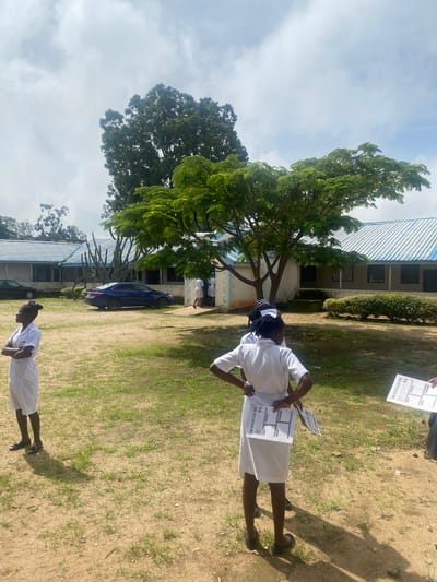 In Dutsen Kato, Nigeria, a group of young women in white uniforms stand outside a building. The building has a light blue, corrugated metal roof and appears to be a single-story structure with multiple windows. In the foreground, two young women are visible, one facing left with her arms crossed and the other facing away from the camera, holding papers. Behind them, other individuals in similar uniforms are seen near the entrance of the building. A blue sedan is parked in front of the building, and several trees, including a large one with a full canopy, are present in the background. The ground is a mix of dry soil and sparse green grass. The sky is partly cloudy.