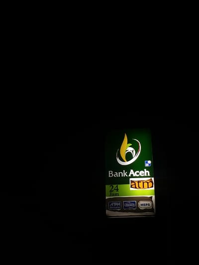 A Bank Aceh ATM sign is visible in the darkness. The sign features a green background, a golden curved logo, and the words "Bank Aceh." Below, it reads "24 Jam" and displays the word "atm." The ATM sign also shows logos for ATM Bersama, Prima, and MEPS network. The sign is illuminated, contrasting with the dark surroundings.
