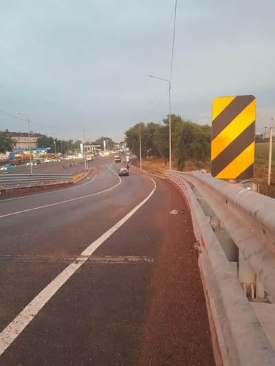 The image shows a highway with traffic and a yellow and black chevron sign indicating a sharp curve ahead. The sky is overcast, suggesting it's either early morning or late evening. The road has lane markings, and there's a guardrail on the right side of the image. In the background, there are buildings and trees, typical of an urban or suburban environment. The traffic appears to be moderate, with several cars visible on the highway. The overall scene is a snapshot of everyday life on a road in the city of Бұлақты, Kazakhstan.
