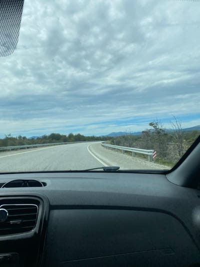 From a car's interior view, a winding road is seen ahead. The road has a white line marker and a silver guardrail along its side, bordered by trees. Beyond the trees are distant mountains. The sky, occupying a significant portion of the image, is filled with dense, layered clouds. The car's dashboard is visible, contrasting with the bright outdoor scene. The windshield wiper is positioned in the middle of the glass.