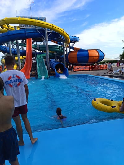 In Yarovoye, Russia, a bright blue pool at a water park is filled with people enjoying a summer day. Several water slides tower over the pool, with colorful tubes in yellow, blue, and orange. One slide features a large, orange and blue spherical section. A lifeguard, wearing a white t-shirt with "СПАСАТЕЛЬ" (Rescuer) and an anchor emblem, stands at the edge of the pool. A child is visible swimming in the pool, and a yellow inner tube floats nearby. In the background, other people are seen relaxing on lounge chairs under a cloudy blue sky. The scene captures the lively atmosphere of a recreational water park.