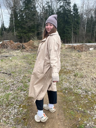 A young woman stands in a natural environment near what appears to be the city of Ivlevo, Russia. She wears a beige trench coat, black pants, white socks, chunky sneakers, and a gray beanie. The environment is primarily dirt and vegetation with trees in the background and a man-made structure visible.