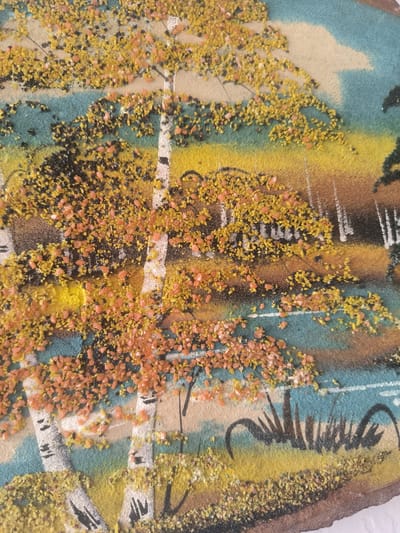 This is a close-up, eye-level shot of a sand art piece depicting an autumnal landscape. The primary subjects are birch trees with their distinctive white bark and black markings. The trees are adorned with clusters of small, granular sand in shades of yellow and orange, representing falling leaves. The background features blended sand textures and colors, creating layers of sky (light blue and pale beige), distant trees with white trunks (suggesting a birch forest), and a body of water or a path in a teal color. The foreground and parts of the middle ground are covered in yellow and brown sand, with dark black sand accents forming grass or reeds along the water's edge. The overall scene evokes a sense of a serene autumn day in a natural setting. There is no visible text in the image. The location context provided is Асқар Тоқпанов ауылы, Kazakhstan.
