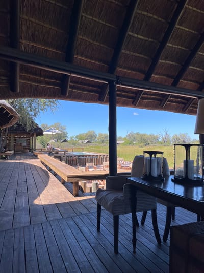 This image was taken from an elevated wooden deck in Khwai, Botswana. The deck is partially covered by a thatched roof supported by wooden beams. A long wooden table with chairs sits on the deck, with lanterns placed as a centerpiece. Beyond the deck, the landscape is visible, featuring grasslands and trees under a clear blue sky. In the distance, structures that appear to be part of a resort or camp are also visible.