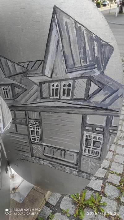 The image is a close-up, eye-level shot of a circular, metallic art installation painted in grayscale. The artwork depicts a building with a distinctive, steeply pitched roof and multiple windows, rendered in a stylized, somewhat abstract manner. The paint strokes are visible, giving the artwork a textured appearance.

The metallic surface of the art piece appears to be mounted on a metal pole, which is bolted to a concrete base. The installation is situated outdoors, with a cobblestone ground visible in the foreground and blurred background elements suggesting a street scene with vehicles and buildings.

Text is visible on the artwork: "TYÖVÄENTALO" is painted horizontally across a section of the building facade, and "SU-26" is inscribed below it. The red watermark "REDMI NOTE 9 PRO AI QUAD CAMERA" and the timestamp "2026/7/8 18:32" are overlaid in the lower portion of the image.

This artwork is located on the exterior of the Workers' House (Työväentalo) in Iisalmi, Finland. The building depicted in the artwork is likely the Iisalmi Workers' House itself, and the "SU-26" could refer to an address, a specific section, or a date related to the building or the artwork. The scene captures a detail of public art within the urban environment of Iisalmi.