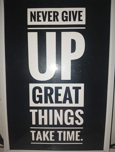 The image shows a framed poster with motivational text. The text is in white, bold, sans-serif font against a black background. The poster reads, "NEVER GIVE UP GREAT THINGS TAKE TIME." The framing is white, and the overall presentation suggests it is a decorative item, possibly in a personal or public space. There is no indication of the city of Ashipa, Nigeria, or any specific location within it. The image is a close-up of the poster, with some dust specks visible on the surface.