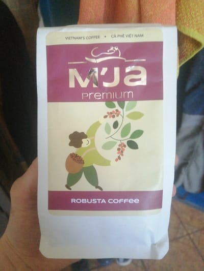 A person's hand is holding a white coffee bag with a maroon and cream-colored label. The label features the brand name "M'ja premium" in silver lettering. Below this, an illustration depicts a simplified figure in a green outfit carrying a basket filled with coffee beans and holding a branch with leaves and berries. The text "ROBUSTA COFFEE" is printed on a maroon band at the bottom of the label. The background is blurry and appears to be indoors, with hints of orange and blue fabric. The image was taken in the city of Achinsk, Russia.