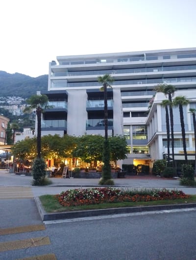 The image captures a street scene in Muralto, Switzerland, featuring a modern, multi-story building with balconies overlooking a street lined with trees and outdoor dining areas. The building's facade is predominantly white with large glass windows and railings. In the foreground, a pedestrian crossing with yellow stripes is visible on the asphalt road. Adjacent to the crossing is a raised flowerbed filled with red, white, and orange flowers. Several palm trees are scattered throughout the scene. An outdoor seating area, illuminated by string lights, is set up in front of the building, with people seated at tables. The background shows a hill with buildings and vegetation. The sky is overcast and the overall lighting suggests it is either early morning or late evening.