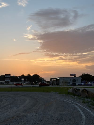 A dusk scene captures a gravel lot in Cardwell, United States, under a sky with wispy clouds tinged with sunset orange and pink. In the foreground, white painted lines delineate a curved path on the gravel. Beyond the lot, a line of trees forms a dark silhouette against the horizon. Several buildings and signs are visible, including a structure with a "CODES" sign and other modular buildings. Cars are parked or driving on a road in the middle ground, and large concrete pipes are stacked along the edge of the gravel area. The overall atmosphere is calm, with the fading light creating a soft, diffused illumination.