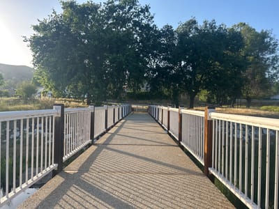 This image depicts a pedestrian bridge in Agoura Hills, California, United States. The bridge features a concrete walkway with white railings on either side, supported by brown posts. The scene is bathed in the warm light of the early morning or late afternoon, casting shadows from the railings across the bridge's surface. The railings are vertical with round horizontal pipes at the top, and the wooden posts are square. Lush green trees dominate the background, with a few houses and a hill visible in the distance. The bridge crosses a grassy, riparian area. The sky is clear and blue.