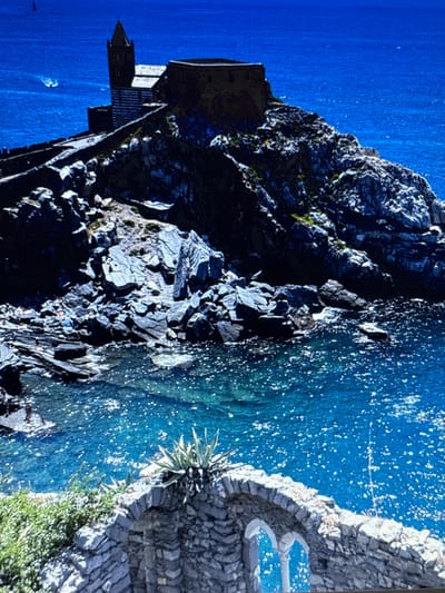 The image shows a striking cliffside scene with a building perched precariously on the edge. The building appears to be made of stone and has a dark, imposing facade. It is situated above a turbulent, deep blue body of water, possibly the sea or a large lake. The water is choppy, with whitecaps indicating some wind or swell. Scattered across the water's surface are numerous bright white specks, possibly reflections of sunlight or spray.
The cliff face is rugged and irregular, composed of dark gray rocks with patches of green vegetation clinging to its surface. The texture of the rocks suggests a natural, weathered formation. On the right side of the image, a stone-built terrace or pathway curves along the edge of the cliff. This structure is made of rough-hewn stones and leads to two distinct, blue-colored shapes that could be decorative pools or seating areas. A spiky, desert-like plant grows from a crevice in the stone wall.
The overall impression is one of dramatic natural beauty and human resilience, with the building clinging to the edge of a formidable landscape. The time of day appears to be daytime, given the bright sunlight reflecting off the water and highlighting the textures of the rocks and the building. The weather seems to be windy, as suggested by the state of the water. The location is identified as Sesto Calende, Italy. There is no visible text in the image.