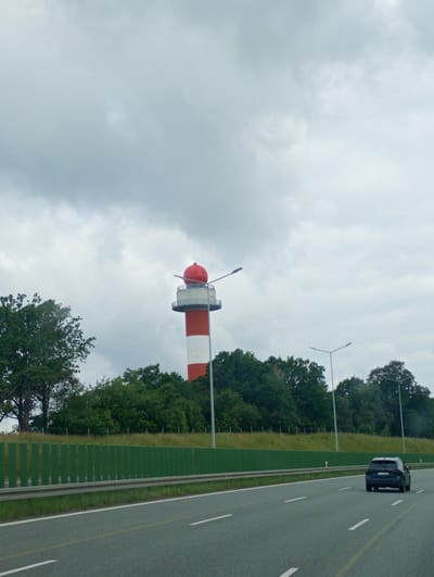 The image captures a view from a highway in Góra Świętej Anny, Poland, looking towards a distinctive red and white tower with a red spherical top, resembling a lighthouse or a similar navigational aid. The tower is situated behind a dense line of green trees. In the foreground, a dark-colored car is driving on the highway, which is marked with white lane lines and a yellow broken line. The sky is overcast with gray clouds. A green noise barrier runs along the side of the highway, separating it from the grassy embankment and trees behind. Several streetlights are visible along the highway.