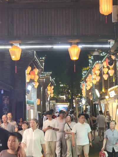 The image depicts a nighttime scene on a street in Wuhou District, China. The street is lined with traditional Chinese architecture, illuminated by numerous orange lanterns. A crowd of people are walking along the street, some looking at shops, others engaged in conversation. In the foreground, a young boy is looking to his left, while an adult woman in a white shirt and a patterned headscarf walks forward. Behind them, a man in a white polo shirt and khaki shorts is looking at his phone. Other individuals are visible further down the street, creating a lively atmosphere. Security cameras are mounted on some of the buildings. The overall impression is of a bustling urban environment at night.