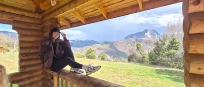 The image captures a young woman perched on a log railing within a wooden structure, likely a small cabin or shelter. The structure is constructed of thick logs and features a rustic, wooden ceiling. The woman wears a dark baseball cap, a puffy brown jacket, black leggings, white socks, and gray and white athletic shoes. She is seated with her legs crossed, her right hand raised to her cap.

Through the opening of the structure, a mountainous landscape is visible. Green hills and valleys are interspersed with trees. In the distance, a taller mountain with rocky peaks is visible under a sky with scattered clouds. The setting suggests a rural or wilderness area.

Given the location is Laki, Bulgaria, the landscape is likely within the Rhodope Mountains.