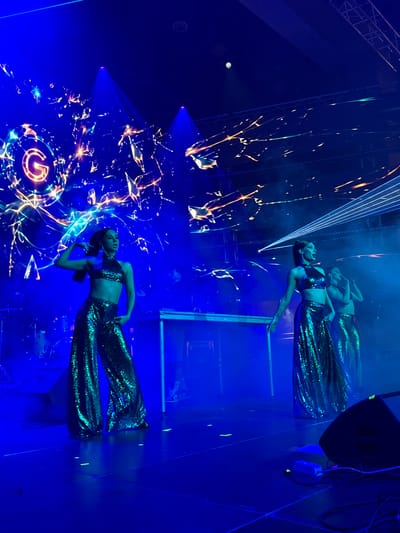 This image depicts a live performance on a stage illuminated by vibrant blue and orange lights. Three female performers, dressed in sequined outfits consisting of halter tops and wide-leg pants, are the central focus. The performer on the left is striking a pose with her arm raised, while the other two are positioned further back. The background features a large screen displaying a graphic with a prominent "G" logo and energetic, abstract designs. Laser lights crisscross the stage from the right side of the frame. Stage equipment, including a DJ console and speakers, is visible. The environment suggests an indoor venue, possibly a concert hall or event space. The location is specified as Akhalubani, Georgia.