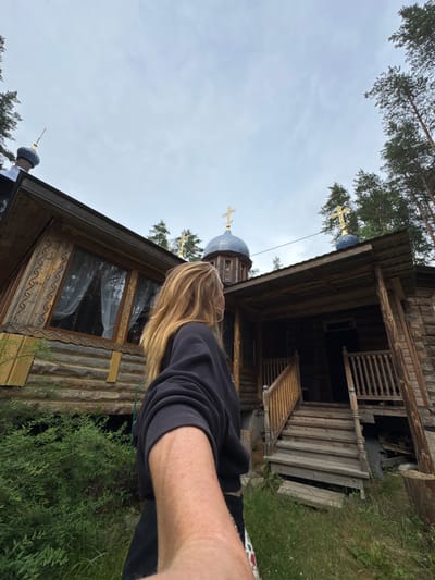 The image shows a person taking a selfie in front of a wooden Orthodox church in Nenimyakii, Russia. The church has a domed roof with a cross on top, and it is surrounded by trees. The person is wearing a black sweatshirt and has long blonde hair. The sky is overcast. The church appears to be made of logs, with intricate carvings around the windows. There is a wooden porch with stairs leading up to the entrance. The area around the church is covered in grass and greenery.
