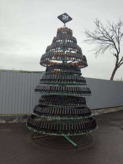 A vertical outdoor shot captures a sculpture in the shape of a Christmas tree made from empty glass bottles in Pskov, Russia. The bottle-tree sculpture features tiered, green metal hoops. Rows of amber and dark-colored bottles encircle each hoop, with the bottle openings facing inward. A black, diamond-shaped sign with red and white designs tops the sculpture. A bare-branched tree stands behind it. The sculpture sits on a gray pavement or asphalt surface next to a gray corrugated metal wall. The sky is overcast.