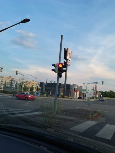 This image shows a street scene in Strakonice, Czechia. The photo is taken from inside a vehicle, looking out at an intersection. The sky is mostly clear with some light clouds, suggesting it is daytime.
The main subjects are traffic signals, a KFC restaurant, and buildings. A traffic light is visible in the foreground, with its red light illuminated. Above the traffic light, a sign with the KFC logo is displayed. In the background, a KFC restaurant building is prominent, with its signature red and white branding. Other buildings, appearing to be apartment blocks or office buildings, are also visible along the street.
A red car is stopped at the intersection, presumably waiting for the traffic light to change. There are no people clearly visible in the image. The setting appears to be a typical urban or suburban environment with roads, sidewalks, and commercial establishments. A pedestrian crossing is marked on the road. A street light is also visible.
The image is rotated 90 degrees counter-clockwise, making the scene appear sideways. There is some reflection on the surface from which the photo is taken, possibly a window. The overall impression is that of a casual observation of a street scene during the day.