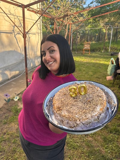 A woman with dark hair and a magenta sweater holds a cake decorated with golden "30" candles in a metal pan. The cake has a textured, crumbly topping. She smiles at the camera. The setting appears to be outdoors in a grassy area in Dolno Linevo, Bulgaria, near trees, an open metal structure, and a concrete surface adorned with decorative flower cutouts.