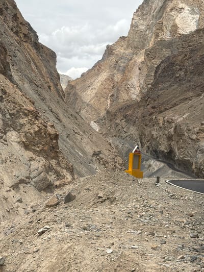A paved road winds through a narrow, rocky mountain pass. The sides of the pass are steep, with exposed rock formations showing layers of sediment and erosion. A bright yellow structure, possibly a signpost or marker, stands beside the road on the right side, topped with a red and white triangular sign. The ground is covered in loose rocks and gravel. The sky is overcast with scattered clouds. This scene is located near Stongde, India, a region characterized by its dramatic mountainous terrain and the presence of such roads.