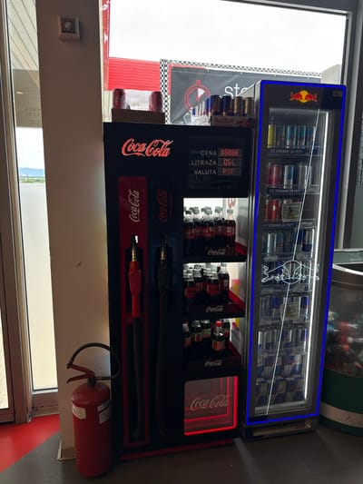 Here's a description of the image:
The image shows an interior view of a commercial space, likely a store or gas station in Cuprija Municipality, Serbia. Dominating the scene is a display unit featuring beverages. The top portion is a refrigerated display case for Red Bull energy drinks. Below it is a Coca-Cola display, designed to resemble old-fashioned gas pumps. The display also features shelves with bottles of Coca-Cola. A fire extinguisher is mounted on the wall next to the Coca-Cola display. A window is visible, showing glimpses of the outside, including what appears to be a parking area and some trees or hills in the distance. The OCR text confirms the presence of Red Bull and Coca-Cola brands. The text includes pricing information in RSD (Serbian Dinar), indicating the location.