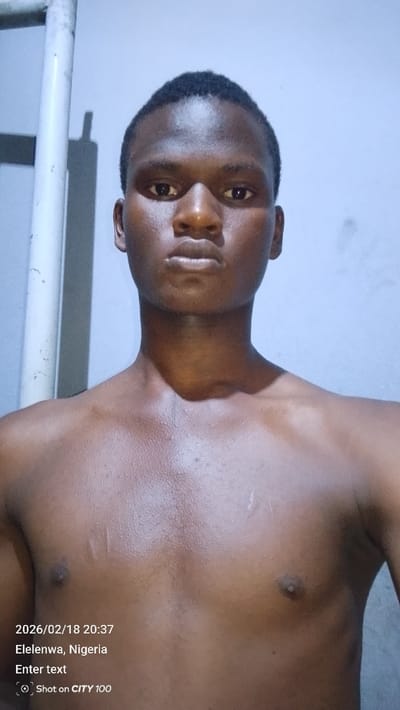 A young, dark-skinned man poses shirtless, facing the camera. His expression is neutral, and his hair is short. The background is a pale wall with a white pole visible on the left. Text overlay includes the date and time "2026/02/18 20:37," location "Elelenwa, Nigeria," and camera details "Shot on CITY 100." The man has visible chest and shoulder muscles.