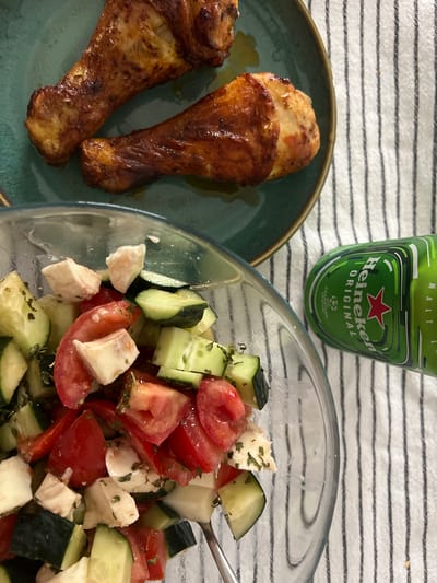 The image depicts a close-up view of a meal consisting of three roasted chicken drumsticks arranged on a dark teal plate, a bowl of chopped salad, and a green Heineken beer bottle. The chicken drumsticks appear golden brown and well-seasoned. The salad in the transparent glass bowl is composed of diced tomatoes, cucumbers, and cubes of white cheese, possibly mozzarella, and is sprinkled with herbs.

In the background, a striped white and black cloth serves as the surface for the meal. The Heineken bottle, with its iconic red star and green label featuring the "Heineken Original" text and Champions League logo, is placed behind the food. The lighting suggests it might be daytime, possibly indoors or outdoors in natural light. The overall presentation is casual, suggesting a meal being enjoyed, perhaps at a picnic or a relaxed home setting. The image does not contain any people, but the arrangement of the food and drink implies an activity of dining or preparing to dine. The visible text on the Heineken bottle clearly identifies the brand and type of beverage. The location context of Mollet del Vallès, Spain, is provided but not visually indicated within the image itself.