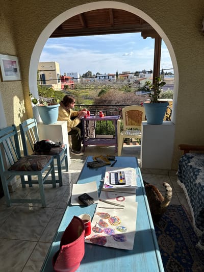 The image depicts a sunlit outdoor or semi-outdoor space in Aegina, Greece, framed by a wide archway with a textured, light yellow stucco wall on the left and a tiled floor. Beyond the arch, a residential area with buildings of varying colors (white, yellow, red) and red-tiled roofs is visible, interspersed with trees and vegetation under a bright, partly cloudy sky.

In the midground, a person is seated at a small, purple metal table positioned near a black metal balcony railing. The individual, identified by light brown hair, is wearing sunglasses, a light brown jacket, dark trousers, and dark sneakers. They are engaged in an activity involving small red objects and two red cups on the table. A light blue potted plant on a white pedestal is situated to the person's left. Two light blue wooden chairs with patterned cushions are positioned to the left foreground; a dark garment with a red element rests on one of the cushions.

In the immediate foreground, a light blue wooden table functions as a workspace. It holds a red fabric container, a red cup, a smartphone, a pair of work gloves, several sheets of paper featuring watercolor paintings (abstract shapes, some resembling fish, and circles), a watercolor palette containing multiple colors, and paintbrushes. A calico or tortoiseshell cat is partially visible on a blue patterned rug to the right of this table, its back and tail in view. Strong sunlight creates prominent shadows across the tiled floor and objects within the scene. A framed picture hangs on the left stucco wall.