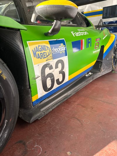 This image depicts a green, blue, and yellow racing car with the number 63 prominently displayed on its side, alongside brand logos such as Pertamina Fastron, Magneti Marelli Checkstar, Dynamic Events, and R Sport. The car features carbon fiber elements and a yellow rearview mirror. The image's location is Campagnano di Roma, Italy, based on the "INDUSTRIAL TRONCA" text, likely referring to an industrial area in the municipality. The car is situated on a reddish-brown surface with circular markings. A black tire is visible in the foreground.