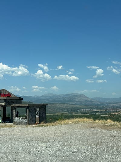 The image depicts a bright, sunny day with a clear blue sky dotted with fluffy white clouds. In the foreground, the ruins of a concrete structure, heavily adorned with graffiti, stand against a backdrop of rugged mountains in the distance. A gravel path runs alongside the ruins, leading the eye towards the expansive landscape. The scene is set in Gornji Kokoti, Montenegro, and suggests an abandoned or unfinished construction site. The graffiti on the ruins indicates it's a place that has seen human activity, possibly by urban explorers or local youths. The time of day appears to be midday or early afternoon, given the strong sunlight casting shadows. The overall mood is one of desolation and decay juxtaposed with the natural beauty of the mountainous terrain and the vibrant blue sky. The graffiti includes various tags and artwork, some legible, such as "BOKI," "15.11.," and "NIKS." There is also a faint silhouette of a person within the structure, adding a touch of mystery and hinting at recent presence.