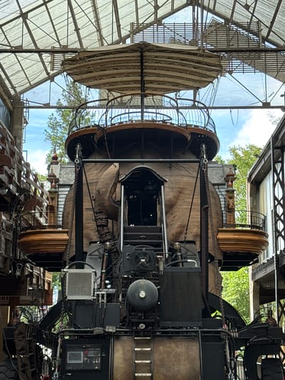 This is a full-frame, eye-level, medium shot of a large, steampunk-inspired mechanical structure resembling a gigantic elephant head, housed within a greenhouse-like structure. The structure is made of dark wood and metal, with intricate details like chains, pipes, and mechanical components visible. The elephant's head has large, wooden tusks and a small, dark entranceway serving as its mouth. Above the head, a tiered platform with a circular railing is suspended by chains, topped with a striped fabric canopy. The entire structure is set against a backdrop of lush green trees and a blue sky, visible through the transparent roof and walls of the greenhouse. The scene is located at Les Machines de l'Île in Nantes, France, a mechanical art park. There are no people visible in the image.