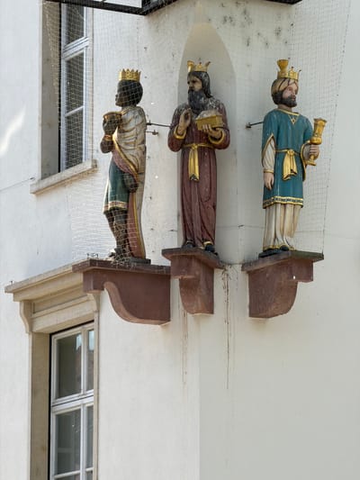 This media file features three carved, wooden figures, each depicting a king wearing a crown and ornate robes. They are mounted on the exterior of a building, appearing to be suspended or attached to a white wall. Each figure is posed in a three-quarter view, with their bodies angled slightly away from the viewer. The figures are richly colored with teal, maroon, and gold accents. The first figure from the top is dressed in teal and gold, holding a golden trumpet-like object. The middle figure wears maroon and gold and holds a golden book or tablet. The bottom figure is adorned in gold and green and holds their hands in front of them.

The figures are positioned vertically in the image, suggesting the photo was taken from below or the scene is oriented vertically. They are protected by a fine, diamond-patterned netting, likely to prevent damage from birds or debris. The figures are mounted on dark brown, rectangular wooden brackets that extend from the wall.

In the background, a portion of a building with white walls and windows is visible. The windows appear to be multi-paned and have light-colored frames. The overall scene suggests an outdoor setting, possibly a public space or the facade of a historical building. There is no visible text in the image. The lighting suggests it could be daytime, with diffuse light. The figures themselves are detailed, showing carved facial features and fabric folds, indicating craftsmanship. The netting and some dust or cobwebs on the brackets suggest the figures are an outdoor installation that may not be frequently cleaned. The location context of Lörrach, Germany, points to a European setting.