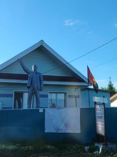 A statue of Vladimir Lenin stands atop a blue fence in front of a light blue house in Kryukovo, Russia. The house has a gray A-frame roof with dark brown trim. A red flag hangs from the side of the house, near the right edge of the building. The fence also features a large blank white banner and a sign that reads "MEA" in large red letters, followed by several lines of Cyrillic text. Vegetation is visible in the foreground. The sky is light blue with a few wispy clouds. Power lines run across the upper right portion of the frame.