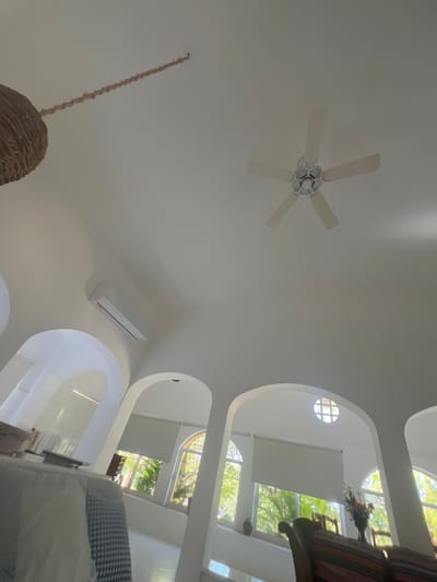 An upward angle shot captures a bright, white interior. A five-blade ceiling fan is centrally positioned with a wicker pendant lamp hanging lower. An air conditioning unit sits near the base of the frame. Arched openings reveal a multi-story design, with glimpses of trees outside through the windows. Furniture partially visible in the foreground and background adds depth. The scene has a clean and modern feel.
