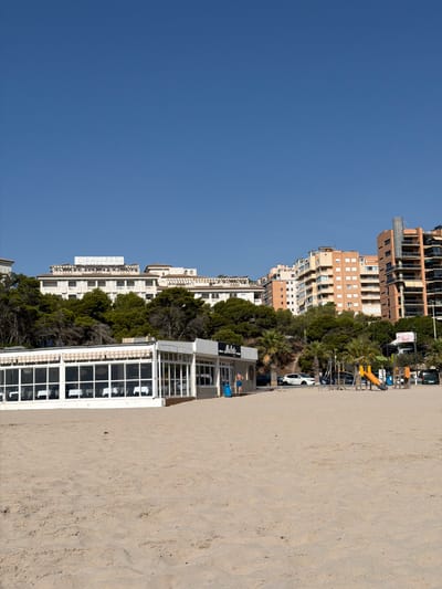 The image depicts a sunlit beach scene with a clear blue sky. In the foreground, there is a vast expanse of sand. Behind the sand, a white building with large windows, appearing to be a restaurant or cafe, is visible. The building has striped awnings. Further in the background, a line of trees, primarily pine trees, separates the beach area from a cluster of multi-story buildings, some of which are hotels. These buildings exhibit a mix of architectural styles and colors, predominantly light beige and orange. To the right of the white building, a small playground with a yellow slide and other play equipment is present. Several cars are parked on a paved area behind the playground and near the buildings. A few individuals are visible: one person in blue shorts stands outside the white building, and two other individuals are walking near the playground area. The overall setting suggests a tourist-oriented location in a coastal city. The signage on the white building partially visible reads "Mallorca".