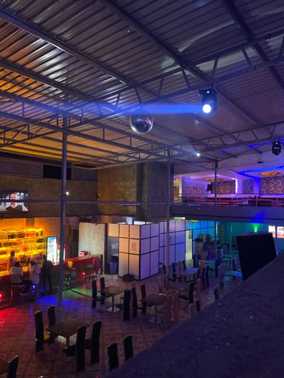 An interior shot captures a lively bar, club, or lounge in Kimorori, Kenya, at night. The scene is viewed from an elevated perspective, looking down across the venue.
The most striking feature is the high, pitched ceiling, constructed from corrugated metal roofing supported by an extensive network of exposed metal trusses and beams. From these structures, a bright blue spotlight shines directly onto a silver disco ball, creating a prominent blue light beam that cuts through the dim ambient light. Another section of the ceiling features decorative panels illuminated with blue and purple accent lighting.
Below the ceiling, the floor is covered with a patterned surface, possibly carpet or tiles. Numerous square-topped tables with dark chairs are scattered across the space, some unoccupied. White, cube-like partition walls are visible, with one displaying a small red square containing illegible white text.
Towards the bottom right, a brightly lit bar counter serves as a focal point. Many bottles are neatly arranged behind the counter, glowing with warm yellow light from below. Several people are seated on stools at the bar, appearing to socialize. One individual is notably wearing a white garment. A large screen near the bar displays an image of a person, but any accompanying text is unclear.
The overall atmosphere is vibrant and social, indicative of an entertainment venue during evening hours, with various colored lights contributing to the dynamic ambiance.