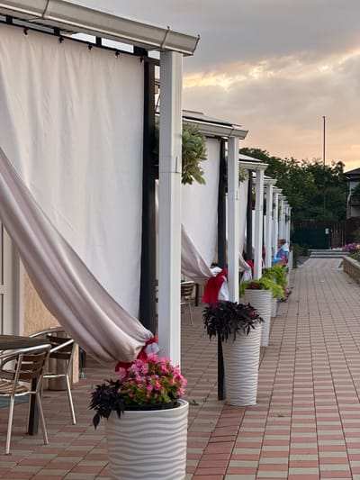 An outdoor dining area with white fabric curtains hanging from a covered walkway is depicted in Yaroslavskaya, Russia. The walkway is lined with white planters containing pink and dark foliage plants. A tiled pathway, alternating pink and beige, extends along the right side of the walkway. In the background, a person is visible standing near the end of the walkway, and beyond that, trees and a building are seen. The sky above is cloudy with hues of orange and yellow, suggesting either sunrise or sunset. The scene appears to be a quiet, possibly evening setting.