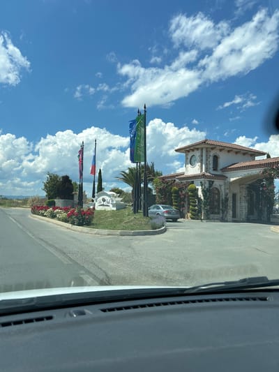 A bright, sunny day is depicted in Tankovo, Bulgaria, with a view from inside a vehicle. The foreground shows the dashboard and windshield of the car. Beyond the windshield, a road leads towards a complex entrance. This entrance features a white arched monument with the words "Elen Park" inscribed on it, flanked by a variety of flags, including the Union Jack, the Russian flag, and a blue and green flag. A silver sedan is parked to the left of the entrance. The building itself has a Tuscan architectural style with a red-tiled roof, stone accents, and archways, adorned with climbing pink roses. The surrounding landscape includes neatly trimmed bushes, flower beds with red and white flowers, and palm trees. The background reveals a distant view of hills under a bright blue sky filled with fluffy white clouds.