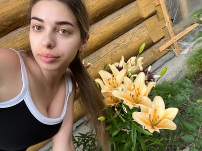 The media file shows a young woman lying on a wooden surface, with her head near the bottom of the frame and her body extending upwards. She is wearing a black tank top with white trim. Her long, brown hair is spread out around her. To her left, a cluster of light orange lilies with dark speckles are in bloom, with green leaves and buds also visible. The wooden surface appears to be made of logs, and there is some transparent plastic sheeting and wooden supports visible on the ground beneath the logs. The scene appears to be outdoors, possibly in a garden or backyard, with natural lighting suggesting daytime. The woman's expression is neutral. There is no visible text in the media file. The provided location context is Ust-Chorna, Ukraine.