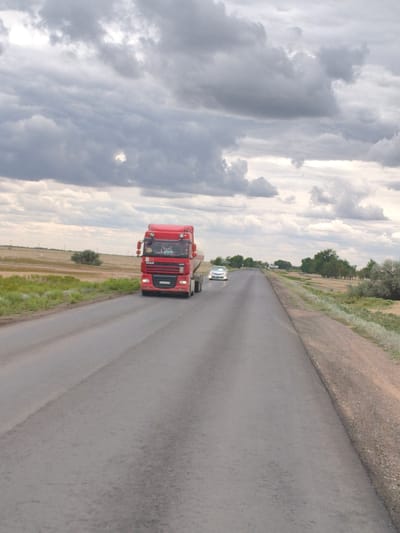A red semi-truck with a white tanker trailer is driving down a paved road in Бударин ауылдық округі, Kazakhstan. The truck is in the foreground, and a white sedan is following behind it. The road is flanked by dry grass and some scattered trees. The sky is overcast with dark clouds.