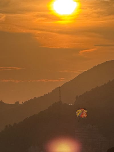 The image captures a serene sunset over Vrijesno, Montenegro. The sky is ablaze with warm orange and yellow hues, with the sun a prominent, bright orb at the top of the frame. Faint clouds streak across the sky, also illuminated by the setting sun. In the mid-ground, layered mountain ranges are silhouetted against the vibrant sky, their slopes covered in dense vegetation. A solitary parasail, adorned with a colorful pattern of red, yellow, and blue segments, drifts gently in the air between two of the mountain ridges, adding a touch of human activity to the natural landscape. The lower portion of the image shows more mountain slopes, with a subtle reflection of the sun's light near the bottom edge, suggesting the presence of water. The overall impression is one of tranquility and natural beauty.