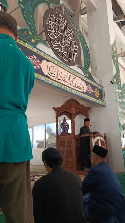 The image depicts the interior of a mosque, likely in Sumedang, Indonesia, during a religious gathering. In the foreground, from left to right, are three individuals facing away from the camera. The person on the far left wears a teal shirt and brown pants. The middle person is seated, wearing a black shirt and a black cap. The person on the right is also seated, wearing a dark blue shirt and a black peci hat, with glasses.
In the midground, a man in a black outfit and peci hat stands at a wooden podium, speaking into a microphone. The podium is ornately carved with geometric patterns and features a decorative wooden canopy above. Behind the podium, a large, dark, oval plaque with white Arabic calligraphy is prominently displayed on the wall. This calligraphy is surrounded by decorative green motifs with small red and yellow floral accents.
The background shows a bright, open space with windows and white tiled walls. The floor is covered with prayer mats in various colors. The overall atmosphere suggests a solemn religious occasion within a mosque.