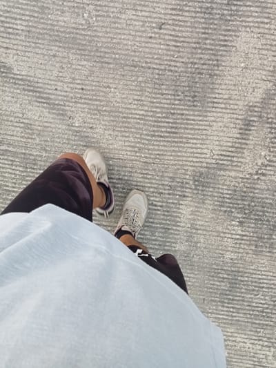 This is a first-person perspective image taken from a high angle, looking down at the legs and feet of a person standing on a textured concrete surface. The concrete has a pattern of parallel lines, typical of a sidewalk or road in an urban environment. The person is wearing dark shorts and a light blue t-shirt that covers part of their legs. They are also wearing athletic shoes, one white with grey accents and the other a lighter grey with white accents. The shoes appear to be slightly worn. The lighting suggests it is daytime, with a slight shadow cast by the person's legs. There is no visible indication of the specific location within Kualu, Indonesia, other than it appears to be an outdoor public space. The image focuses on the lower body of the individual and the ground beneath them.