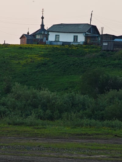 The image depicts a rural scene in Potapovo, Russia, featuring a cluster of buildings on a grassy hill. A prominent white house with a dark, corrugated roof stands centrally. To its left is a dark-colored building with a similar roof and a tall, dark spire topped with a cross, indicative of a church or chapel. Adjacent to this structure is a smaller, wooden outbuilding. Several satellite dishes are visible on the main house and the church building. The hill is covered in lush green grass with scattered patches of yellow wildflowers. In the foreground, a dark, muddy area with some rocks and sparse vegetation suggests a pathway or unpaved ground. The sky is a pale, hazy orange, indicating either dawn or dusk. There are no people or animals clearly visible in the image. Power lines are strung across the upper portion of the scene, supported by wooden poles.